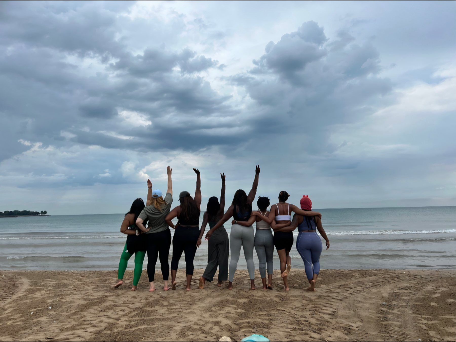 A group of women standing arm in arm on the sand facing the sea, raising their hands together in celebration