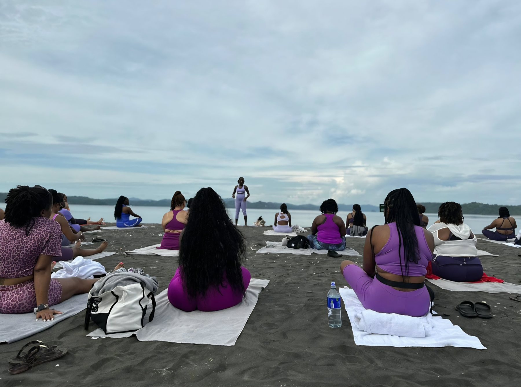 A group seated on towels along a calm shoreline under a soft overcast sky, facing the water in quiet meditation