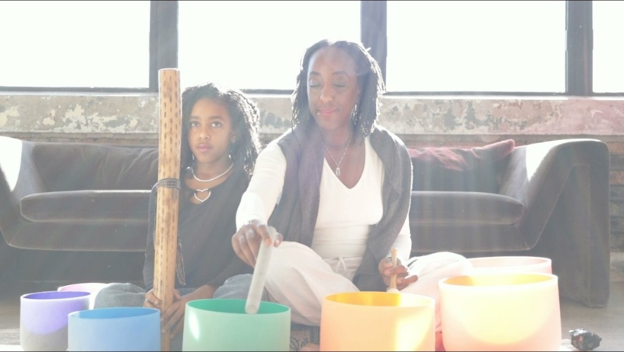 Andrea “Zen Goddess” plays a crystal singing bowl with a mallet while her daughter Ava Rae holds a rainstick beside her, seated among colorful sound bowls in a sunlit studio.