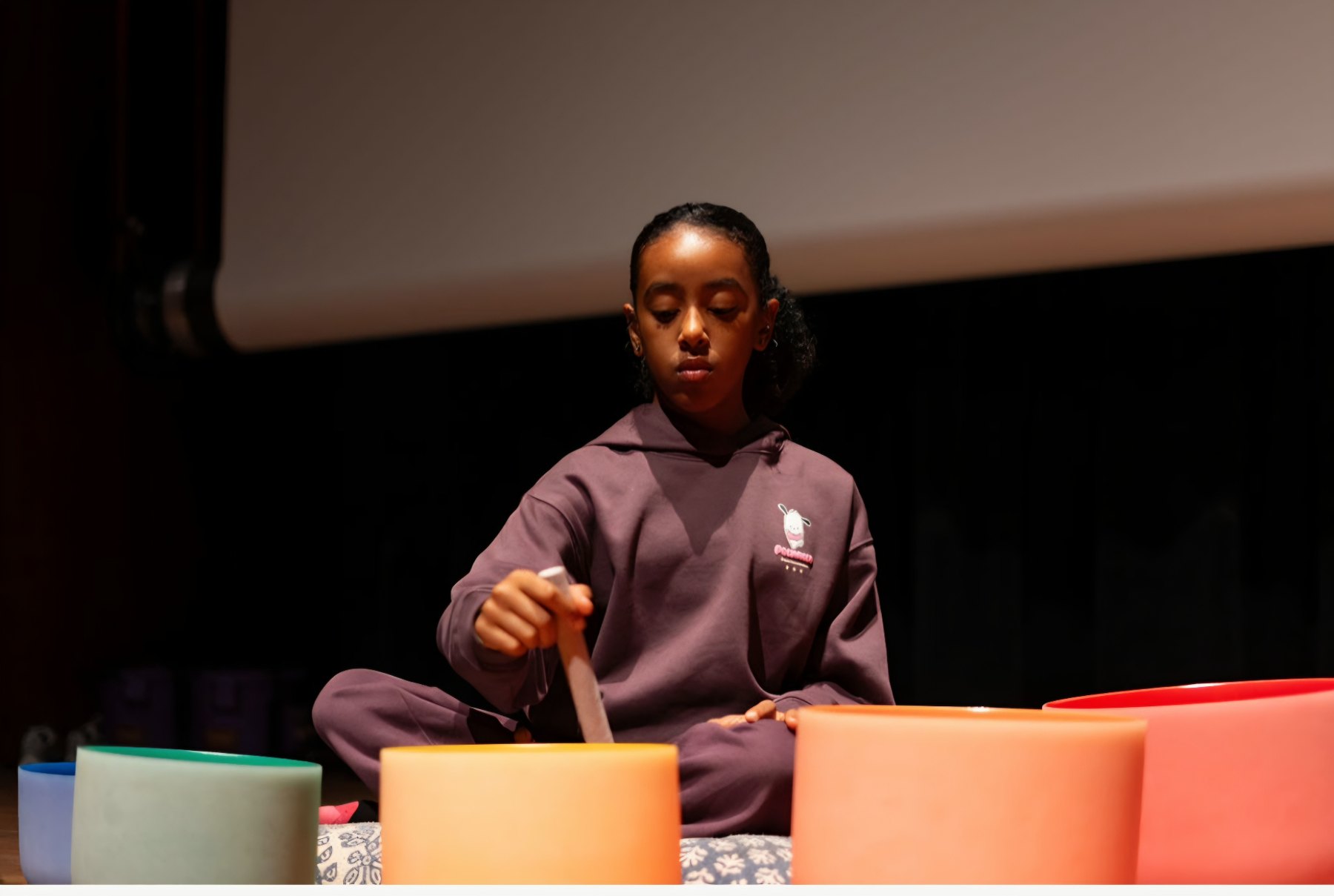 A young girl in a plum hoodie sits on a stage, focused as she plays a row of pastel crystal singing bowls