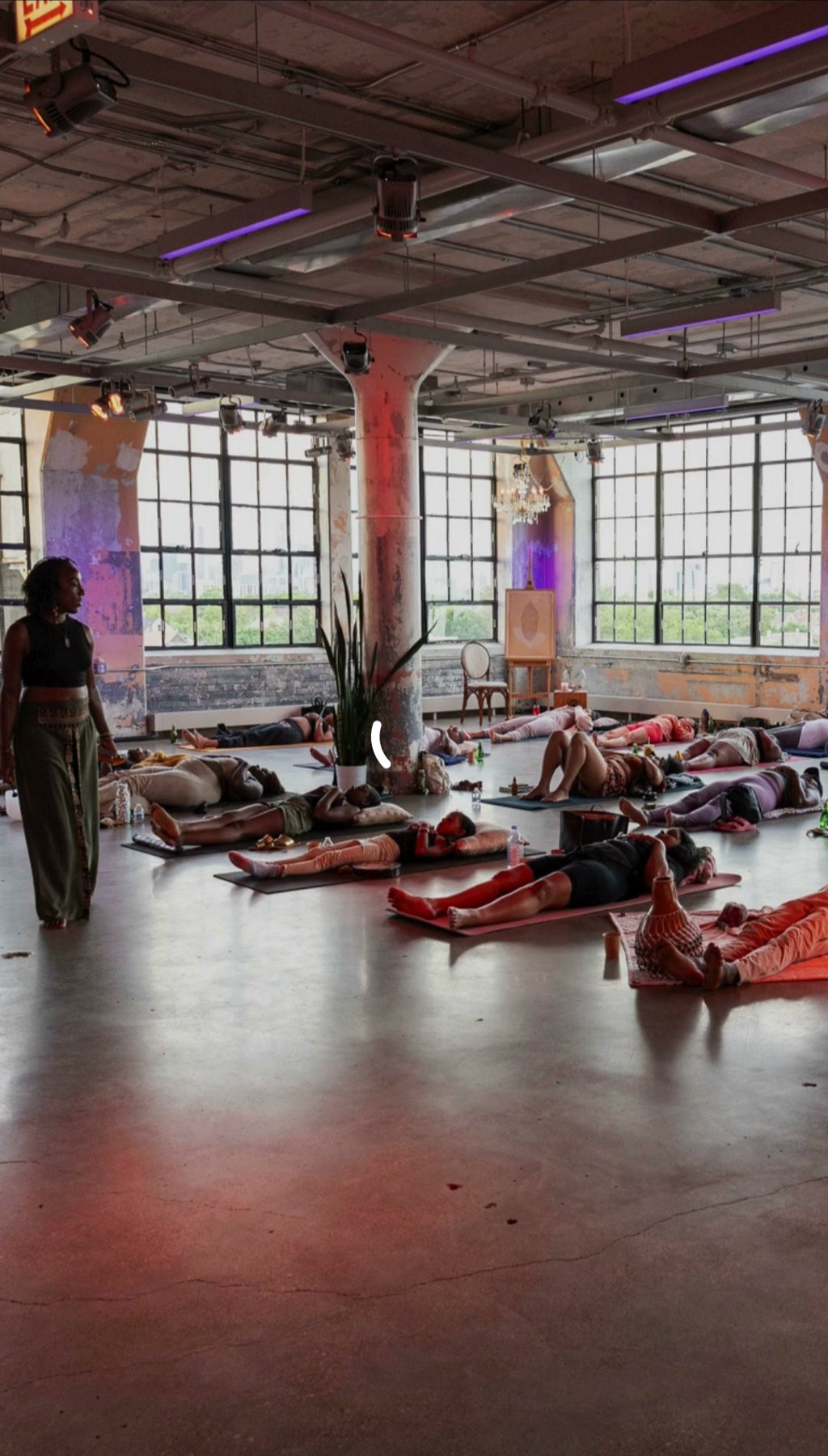 Andrea standing at the edge of a sunlit loft as a large group rests on mats under soft red and purple lighting