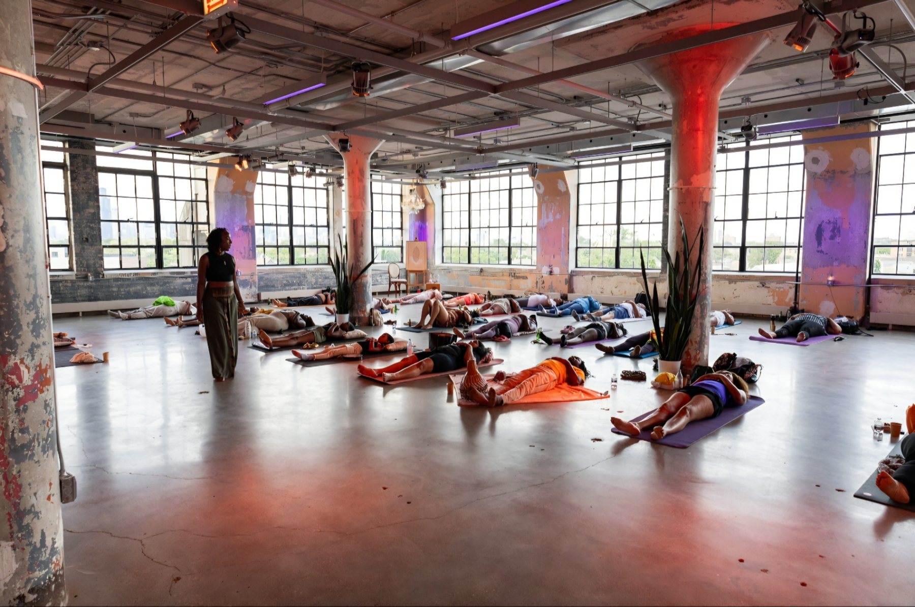 A wide industrial loft with tall windows and warm red and violet lighting, filled with participants resting on mats during a guided meditation