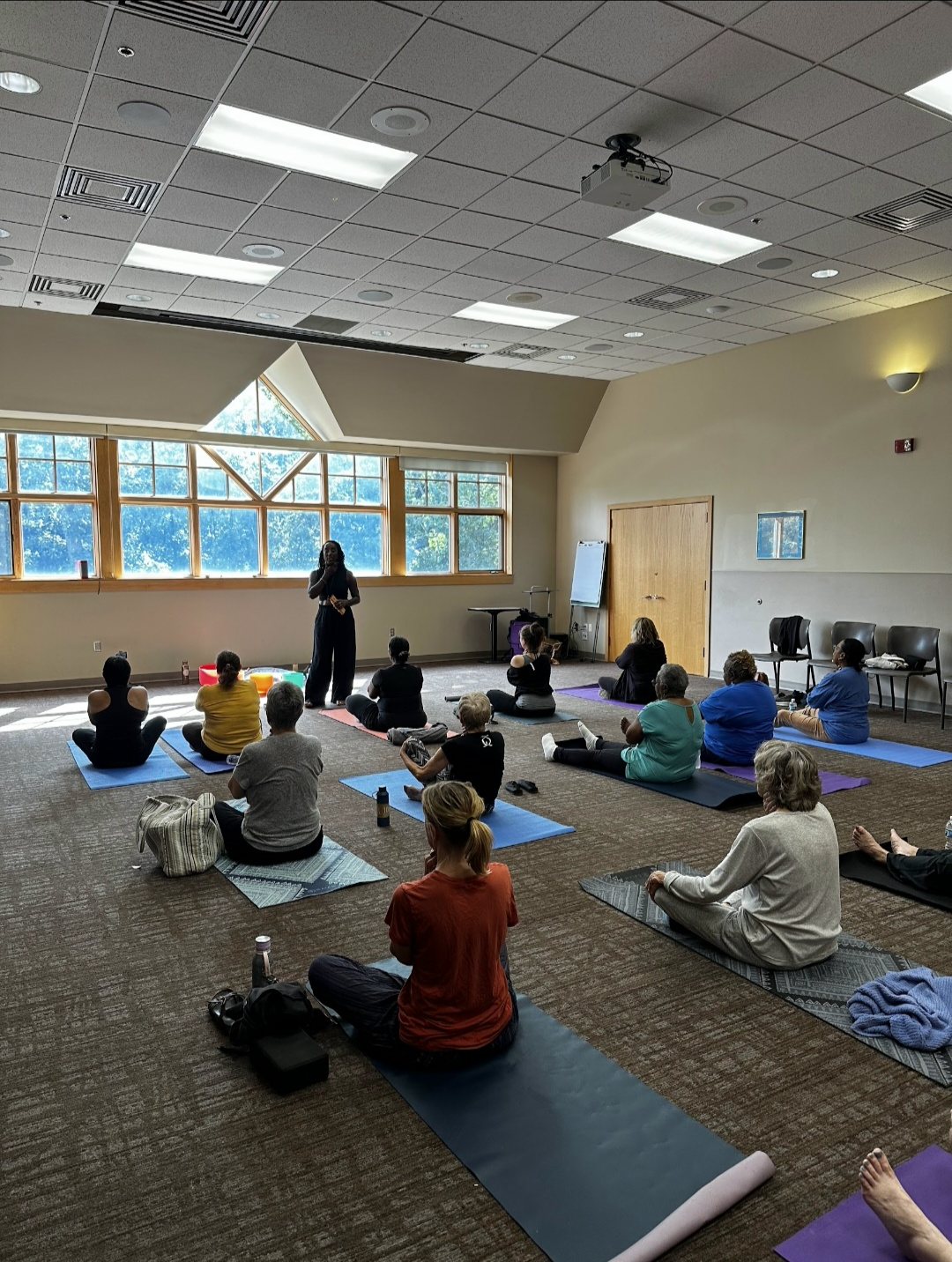 A bright community room with participants seated on yoga mats as Andrea leads a session from the front