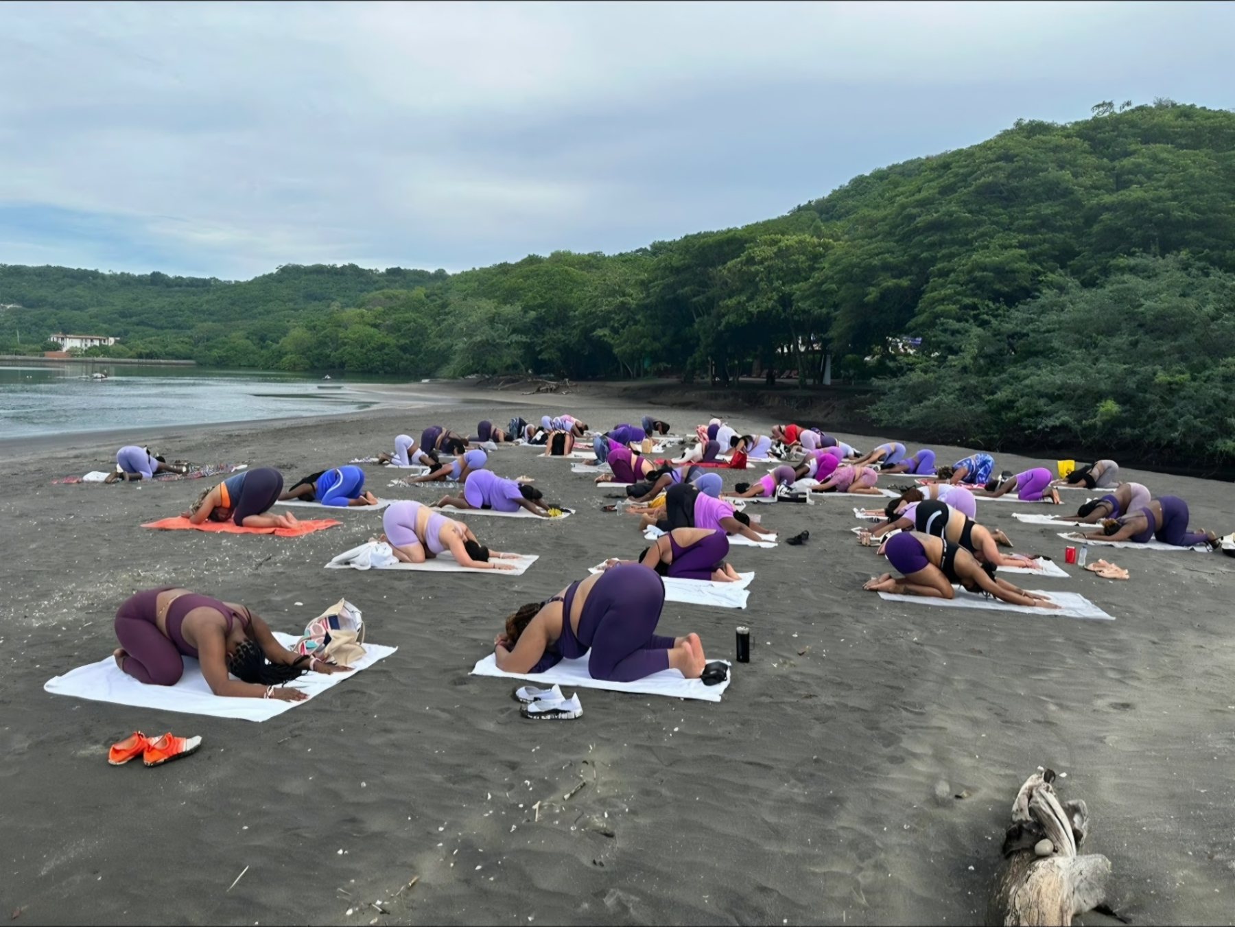 Dozens of participants in a resting pose on mats across a dark-sand beach framed by green forested hills