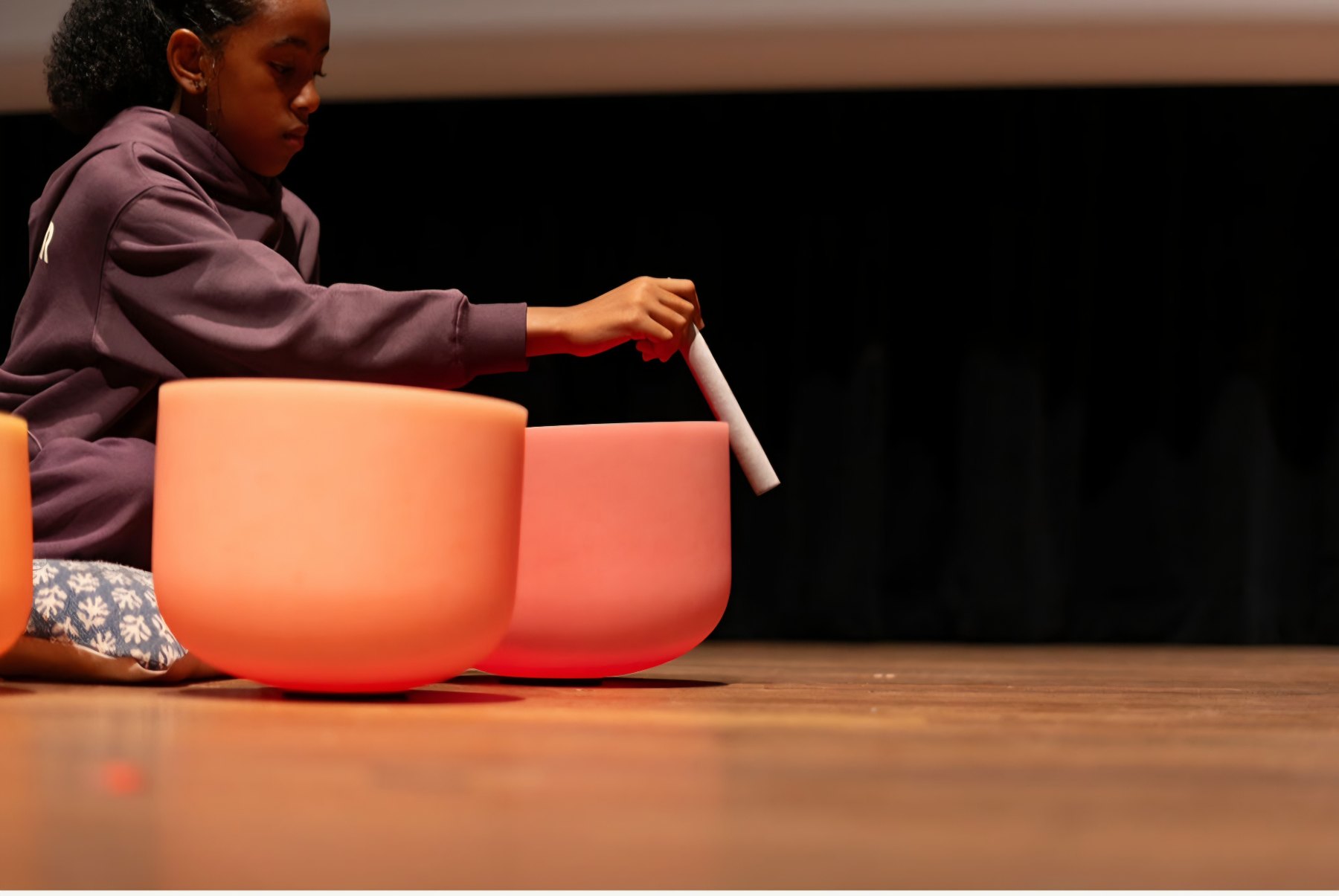 Close view of a young girl drawing a mallet around coral crystal singing bowls on a wooden stage