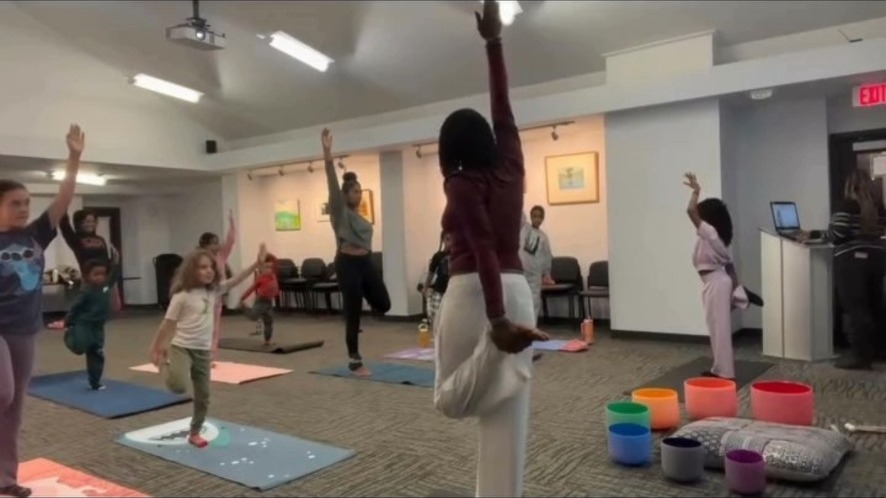 Andrea leads a standing balance-pose class with adults and children practicing together on yoga mats, crystal singing bowls resting in the foreground.