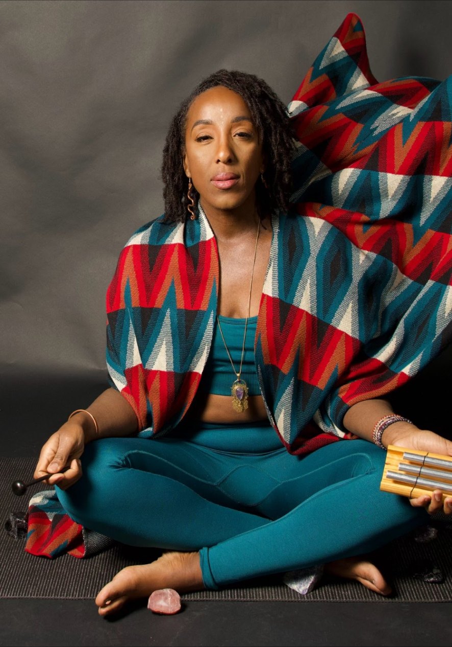 Professional portrait of Andrea seated cross-legged in teal, wrapped in a red, white and blue chevron blanket, holding a chime with a rose quartz crystal at her feet