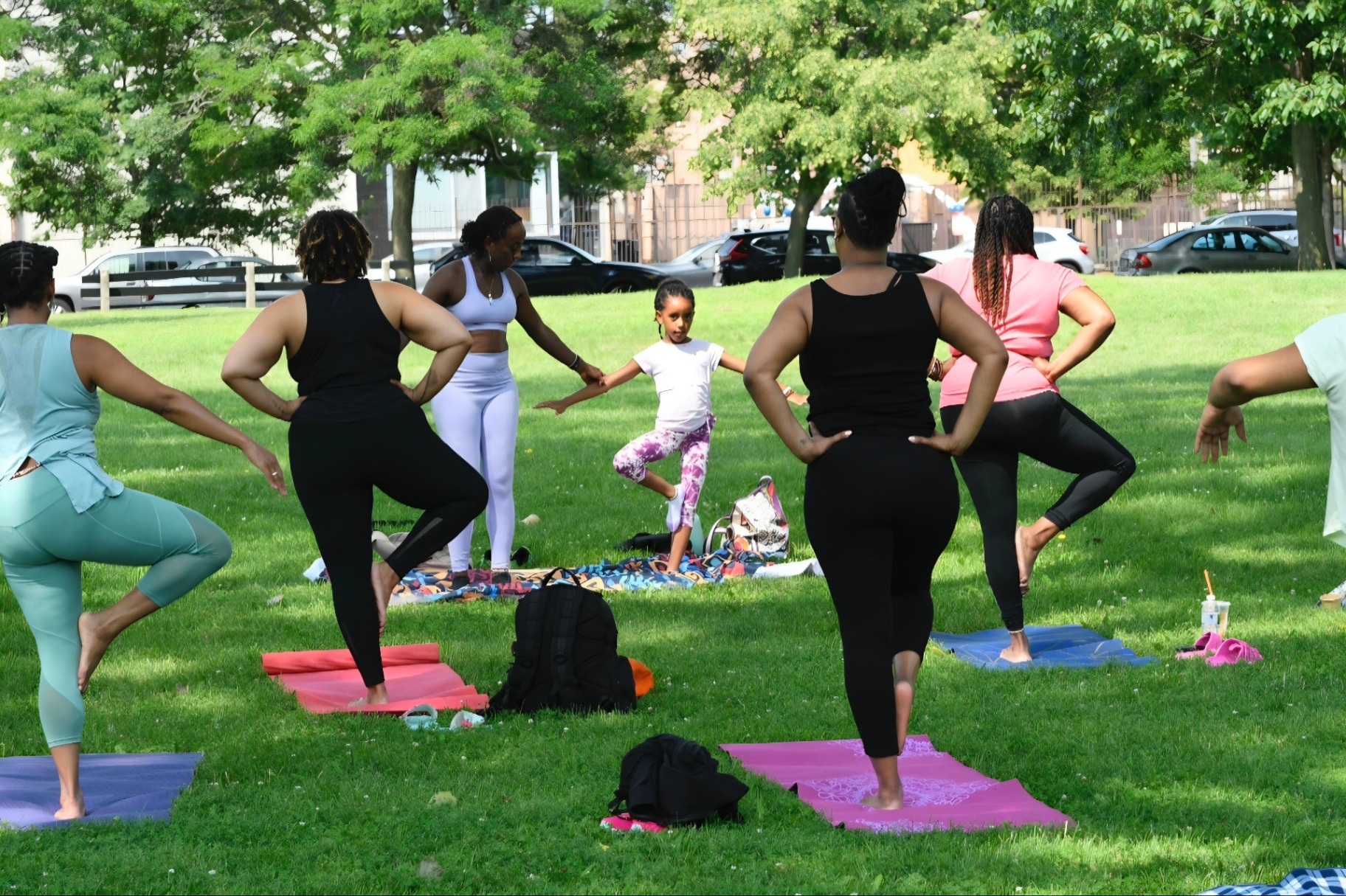 A circle of women and a young girl practicing standing balance poses on colorful mats in a sunny park