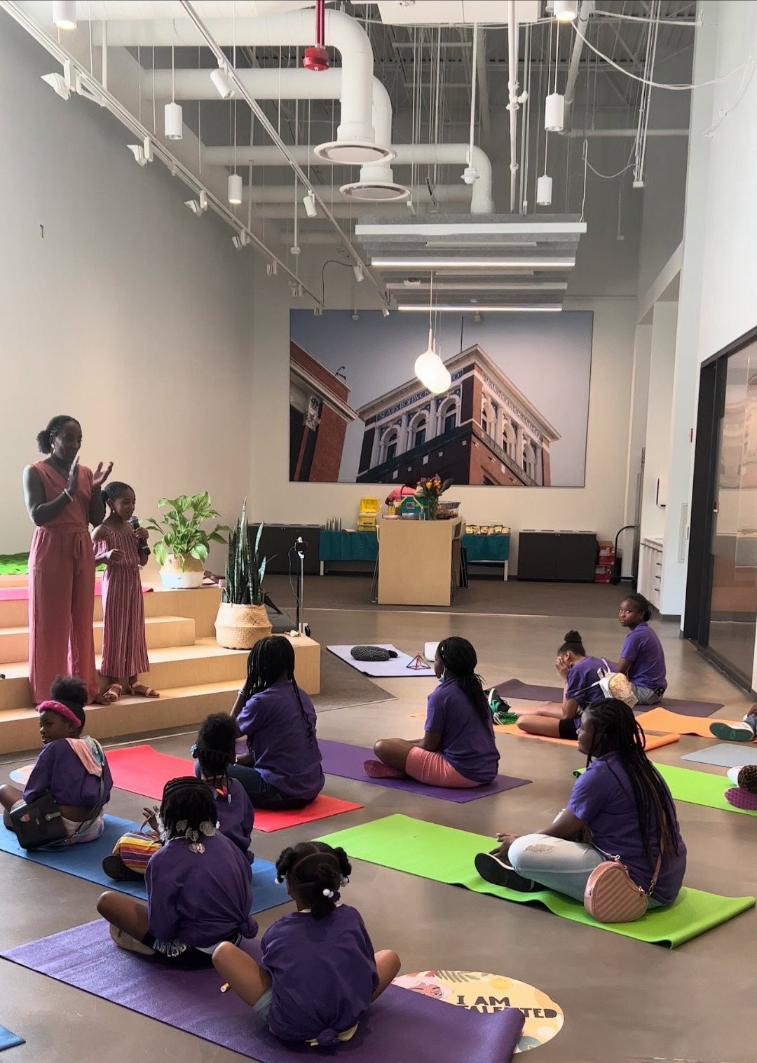 Andrea and Ava leading a mindfulness session for a circle of children seated on colorful yoga mats in a bright loft